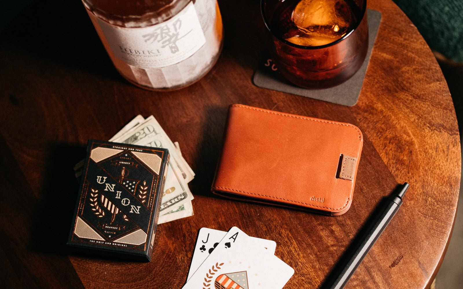 Sitting on a table with playing cards and a glass of Japanese whiskey, Distil Union leather Wally Bifold wallet has evolved since it was launched on Kickstarter in 2014 