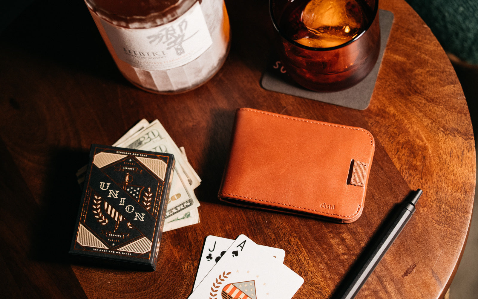 Sitting on a table with playing cards and a glass of Japanese whiskey, Distil Union leather Wally Bifold wallet has evolved since it was launched on Kickstarter in 2014